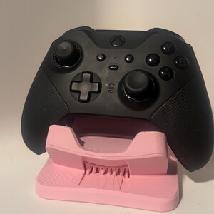 Puede incluir: Un mando negro de Xbox descansa sobre un soporte rosa. El mando tiene botones, joysticks y una cruceta. El soporte es de color rosa claro y tiene una superficie texturizada.