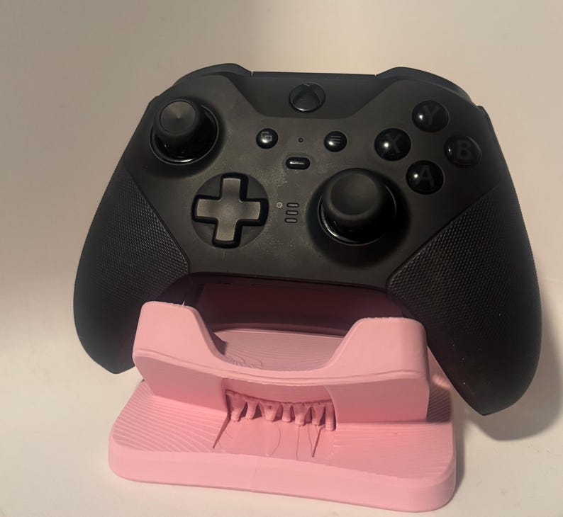 Puede incluir: Un mando negro de Xbox descansa sobre un soporte rosa. El mando tiene botones, joysticks y una cruceta. El soporte es de color rosa claro y tiene una superficie texturizada.