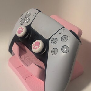 Puede incluir: Un mando de videojuegos blanco y negro con joysticks rosas con huellas de pata descansa sobre un soporte rosa. El mando tiene una variedad de botones y controles direccionales. El soporte tambi&eacute;n es rosa y est&aacute; dise&ntilde;ado para sujetar el mando.