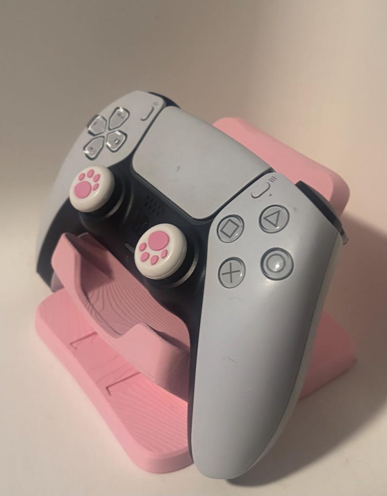 Puede incluir: Un mando de videojuegos blanco y negro con joysticks rosas con huellas de pata descansa sobre un soporte rosa. El mando tiene una variedad de botones y controles direccionales. El soporte tambi&eacute;n es rosa y est&aacute; dise&ntilde;ado para sujetar el mando.