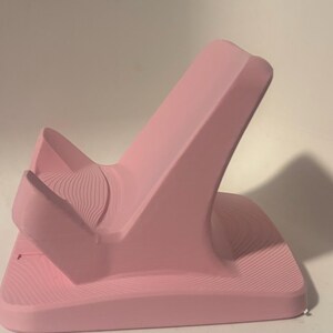 Puede incluir: Un soporte para tel&eacute;fono rosa con un dise&ntilde;o curvo. El soporte tiene una base ancha y una parte trasera alta e inclinada, con una secci&oacute;n curva m&aacute;s peque&ntilde;a en la parte delantera. La superficie tiene un patr&oacute;n texturizado en forma de onda.