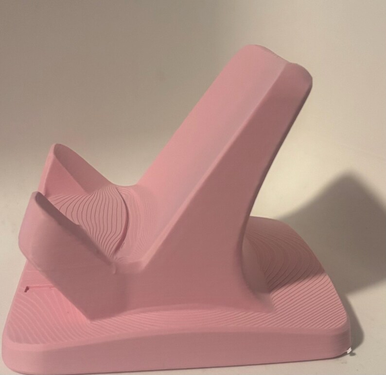 Puede incluir: Un soporte para tel&eacute;fono rosa con un dise&ntilde;o curvo. El soporte tiene una base ancha y una parte trasera alta e inclinada, con una secci&oacute;n curva m&aacute;s peque&ntilde;a en la parte delantera. La superficie tiene un patr&oacute;n texturizado en forma de onda.