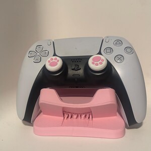 Puede incluir: Un mando de PlayStation 5 blanco y negro con agarres para el pulgar con huellas de pata rosas descansa sobre una estaci&oacute;n de carga rosa. El mando tiene una secci&oacute;n central negra y laterales blancos.