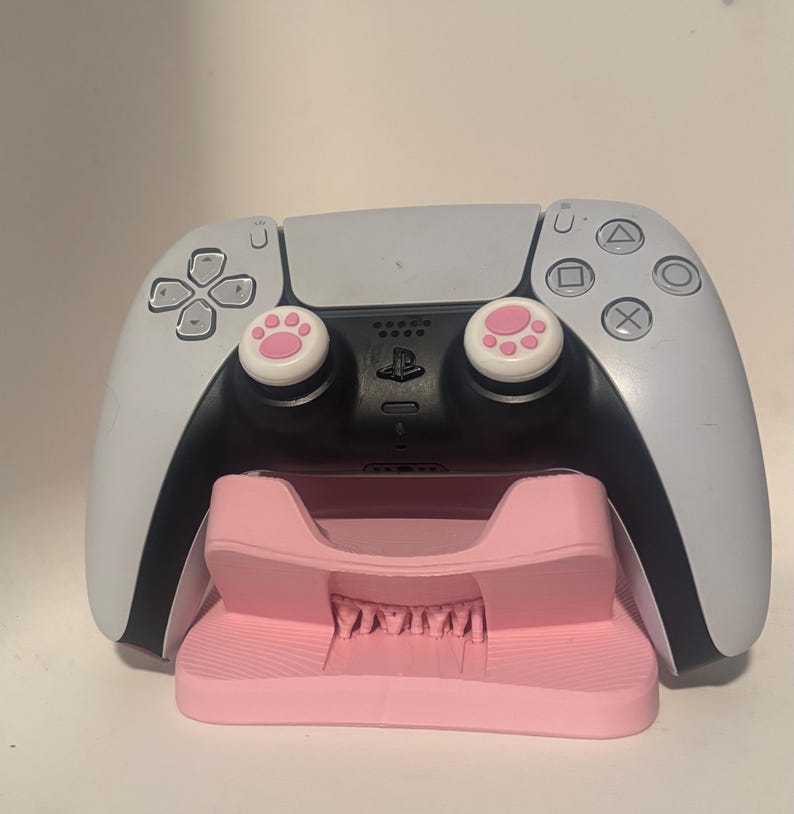 Puede incluir: Un mando de PlayStation 5 blanco y negro con agarres para el pulgar con huellas de pata rosas descansa sobre una estaci&oacute;n de carga rosa. El mando tiene una secci&oacute;n central negra y laterales blancos.