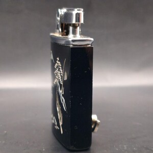 May include: A black rectangular lighter with a silver metal top and a silver floral design. The lighter has a small silver button on the side. The background is a neutral gray.