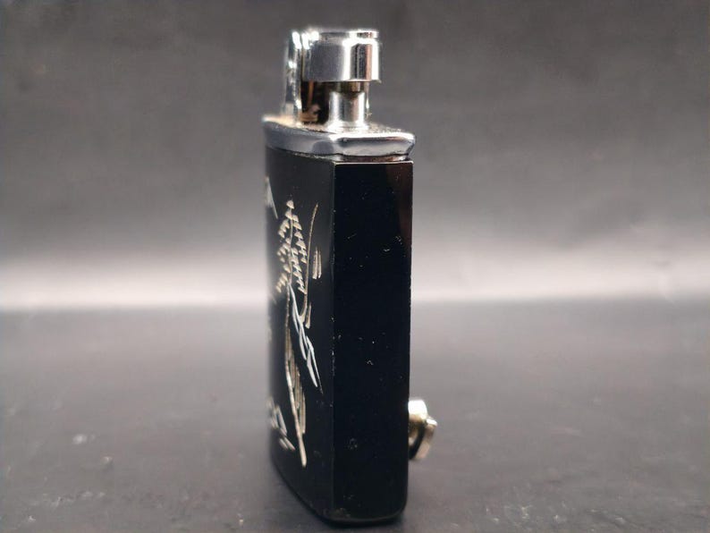 May include: A black rectangular lighter with a silver metal top and a silver floral design. The lighter has a small silver button on the side. The background is a neutral gray.