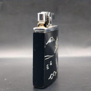 May include: Black rectangular lighter with a silver metal top. The side of the lighter features a mountain scene with Japanese characters in silver. The lighter has a silver metal top and a gold-colored spark wheel.