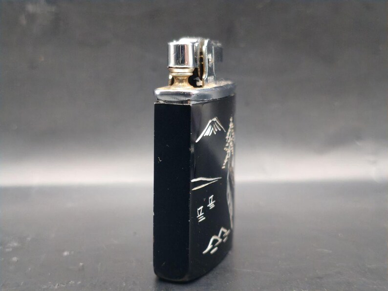 May include: Black rectangular lighter with a silver metal top. The side of the lighter features a mountain scene with Japanese characters in silver. The lighter has a silver metal top and a gold-colored spark wheel.
