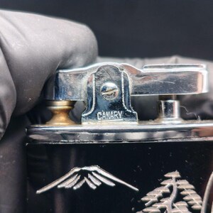 May include: Close-up of a vintage black lighter with a silver-colored top. The word "CANARY" is engraved on the top. The lighter features a mountain and tree design on the front.