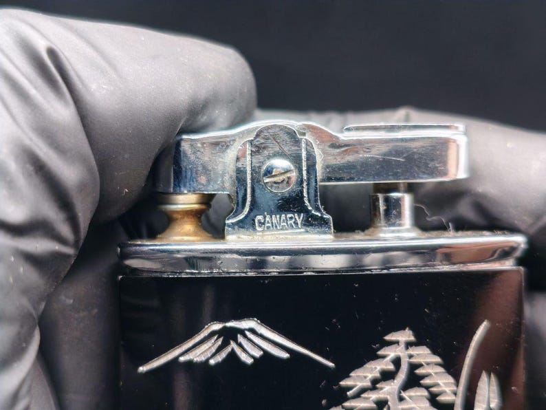 May include: Close-up of a vintage black lighter with a silver-colored top. The word "CANARY" is engraved on the top. The lighter features a mountain and tree design on the front.