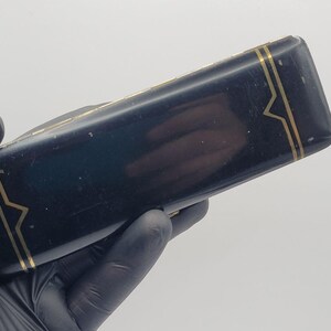 May include: A rectangular black case with a glossy finish and gold-toned linear accents. The design features thin gold lines running along the edges, forming geometric patterns at both ends of the elongated, smooth, and dark-coloured container.