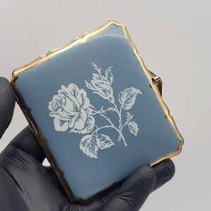 May include: A square compact case featuring a matte dusty blue surface with a white floral rose illustration in the center. The compact is framed by a scalloped gold-tone metal edge with a hinge mechanism visible on the right side.