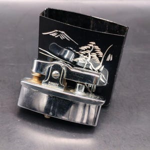 May include: A vintage chrome and gold-toned Canary brand lighter with a black box. The box features a white landscape design with a mountain and tree. The lighter is rectangular with a curved top and the word "CANARY" is visible.