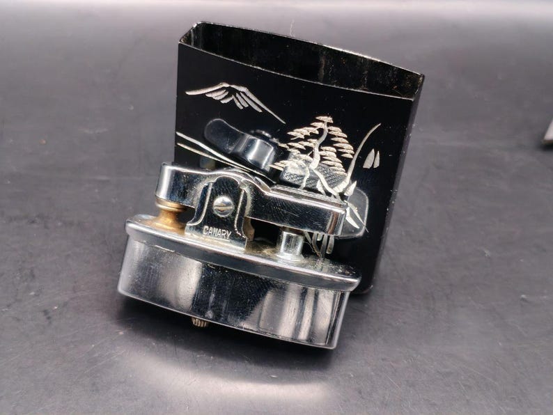 May include: A vintage chrome and gold-toned Canary brand lighter with a black box. The box features a white landscape design with a mountain and tree. The lighter is rectangular with a curved top and the word "CANARY" is visible.
