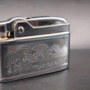 May include: A rectangular silver-tone metal lighter featuring an engraved scene on its front surface. The engraving depicts a landscape with birds flying over a body of water and stylized clouds above. The lighter has a smooth, polished finish.