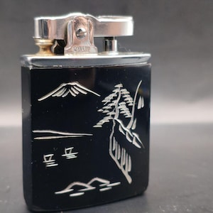 May include: A vintage black lighter with a silver metal top. The lighter features a detailed white etched design of a mountain, trees, and Japanese characters. The lighter is rectangular and has a silver metal hinge.