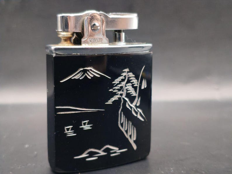 May include: A vintage black lighter with a silver metal top. The lighter features a detailed white etched design of a mountain, trees, and Japanese characters. The lighter is rectangular and has a silver metal hinge.