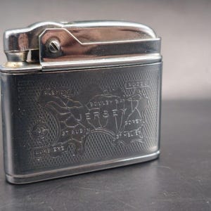 May include: A rectangular silver-tone metal lighter featuring an engraved map of Jersey on its front surface. The map includes labels for locations such as Bouley Bay, St. Aubin, Gorey, St. Helier, Rozel, and Corbiere. The lighter has a hinged top lid.