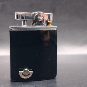 May include: A vintage black lighter with a silver-colored top and a small brass-colored detail. The lighter has a rectangular shape and a rounded bottom with a silver-colored button. The lighter is likely made of metal.