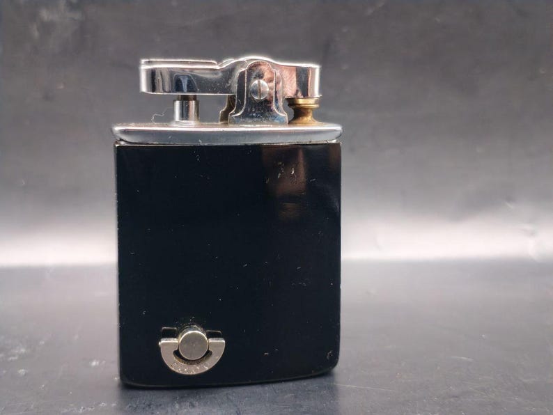 May include: A vintage black lighter with a silver-colored top and a small brass-colored detail. The lighter has a rectangular shape and a rounded bottom with a silver-colored button. The lighter is likely made of metal.