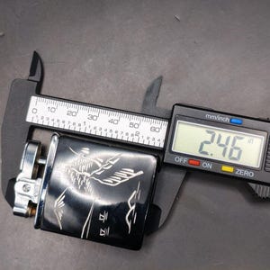 May include: A black lighter with a silver design and a digital caliper measuring 2.46 inches. The lighter has an engraved design. The caliper is black and silver with a digital display.