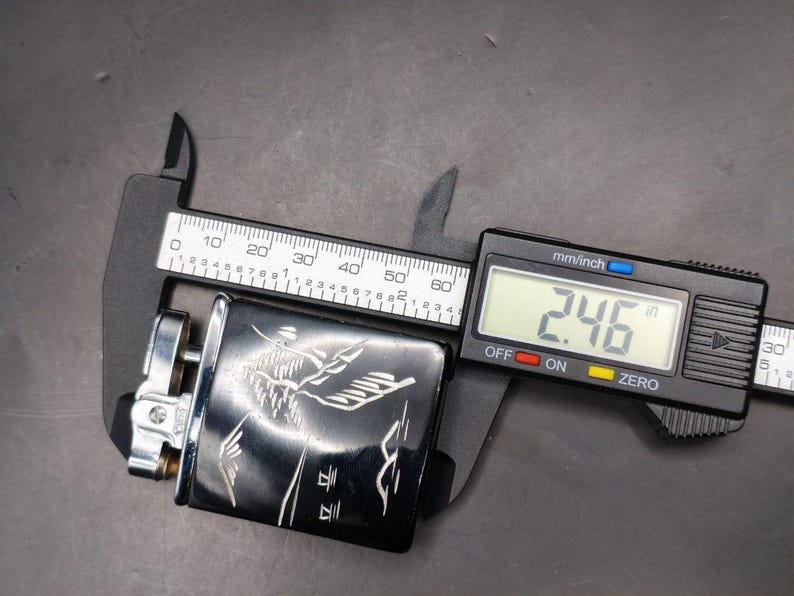 May include: A black lighter with a silver design and a digital caliper measuring 2.46 inches. The lighter has an engraved design. The caliper is black and silver with a digital display.