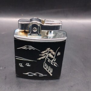 May include: Vintage Canary lighter with a black body and silver accents. The lighter features a Japanese landscape design in white, including a mountain and stylized trees. The top is silver with a textured surface for ignition.