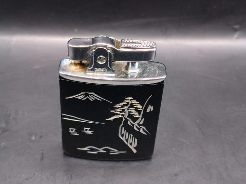 May include: Vintage Canary lighter with a black body and silver accents. The lighter features a Japanese landscape design in white, including a mountain and stylized trees. The top is silver with a textured surface for ignition.