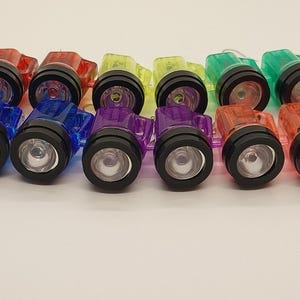 May include: An assortment of colorful mini flashlights in red, blue, purple, yellow, green, and orange. Each flashlight has a black bezel and a clear lens. The flashlights are arranged in a row on a white surface.