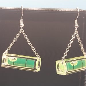 May include: A pair of novelty earrings featuring miniature spirit levels. Each level is encased in clear acrylic with a green liquid bubble and suspended from a silver-colored chain and hook. The earrings are designed to resemble miniature construction tools.