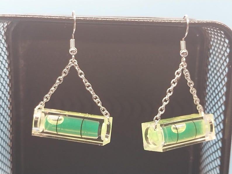May include: A pair of novelty earrings featuring miniature spirit levels. Each level is encased in clear acrylic with a green liquid bubble and suspended from a silver-colored chain and hook. The earrings are designed to resemble miniature construction tools.