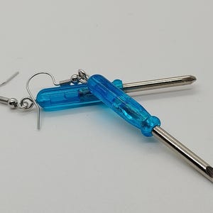 May include: A pair of novelty earrings featuring miniature screwdrivers. Each earring has a bright blue handle and a silver-colored metal shaft. The earrings are designed with a hook-style closure for pierced ears.