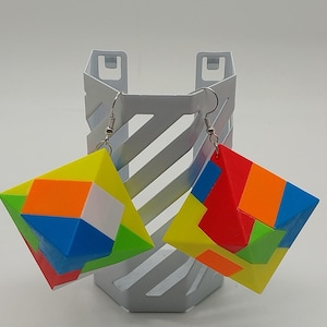 May include: Geometric cube earrings with a colorful, puzzle-like design. The earrings feature a mix of bright colors including yellow, orange, red, blue, and green. They hang from silver-colored ear wires. The earrings are displayed on a white metal stand.