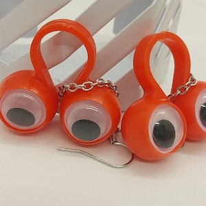 May include: A pair of novelty earrings featuring orange, googly-eye shaped charms. Each charm has a large, white iris with a black pupil. The charms are connected with a silver chain and hang from a silver fish hook.