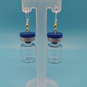 May include: A pair of unique earrings featuring small, clear glass vials with blue caps. The vials are suspended from silver-colored ear wires with gold-colored beads and small heart-shaped charms. The background is a solid light blue.