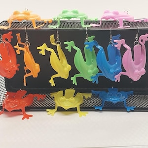 May include: A collection of colorful frog earrings in orange, yellow, green, blue, and pink. The earrings are displayed on a black wire mesh organizer. Some frogs are hanging, while others are standing.