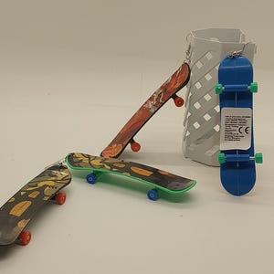 May include: Miniature skateboards in various designs. One is blue with green wheels, another has a camouflage pattern with green trim, and a third features a dragon design. The skateboards have red and blue wheels.
