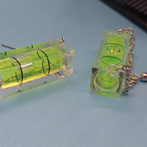 May include: A pair of novelty earrings featuring miniature spirit levels. Each earring has a clear, rectangular body with a vibrant green liquid and a bubble. The earrings are connected by silver-colored chains and hooks.