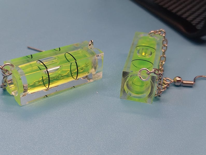 May include: A pair of novelty earrings featuring miniature spirit levels. Each earring has a clear, rectangular body with a vibrant green liquid and a bubble. The earrings are connected by silver-colored chains and hooks.