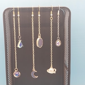 May include: A collection of dangle earrings with celestial-themed charms. The earrings feature gold-colored chains and hooks, with charms including a moon, planet, and teardrop-shaped iridescent crystals. The earrings are displayed in a black wire mesh organizer.