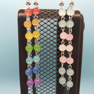 May include: Two pairs of dangle earrings. One pair features a rainbow of colors, including red, orange, yellow, green, blue, and purple beads. The other pair has light pink and blue beads. Both pairs have silver-colored posts and connecting chains.
