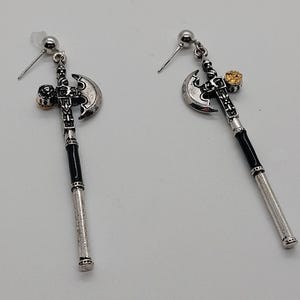 May include: A pair of silver-toned axe-shaped earrings. The earrings feature a black handle, a silver blade, and a small gold-colored accent. The earrings are designed with a post-back closure.