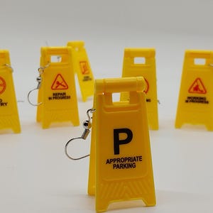 May include: Yellow miniature road sign earrings with various printed warnings, including "No Entry," "Repair in Progress," and "Appropriate Parking." The earrings have silver-colored hooks.