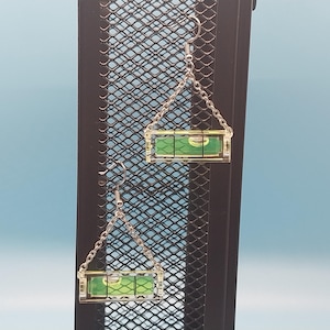 May include: A pair of novelty earrings featuring miniature levels. Each earring has a clear, rectangular level with a green liquid bubble, suspended from a silver chain and hook. The earrings are displayed against a black metal grid with a blue background.