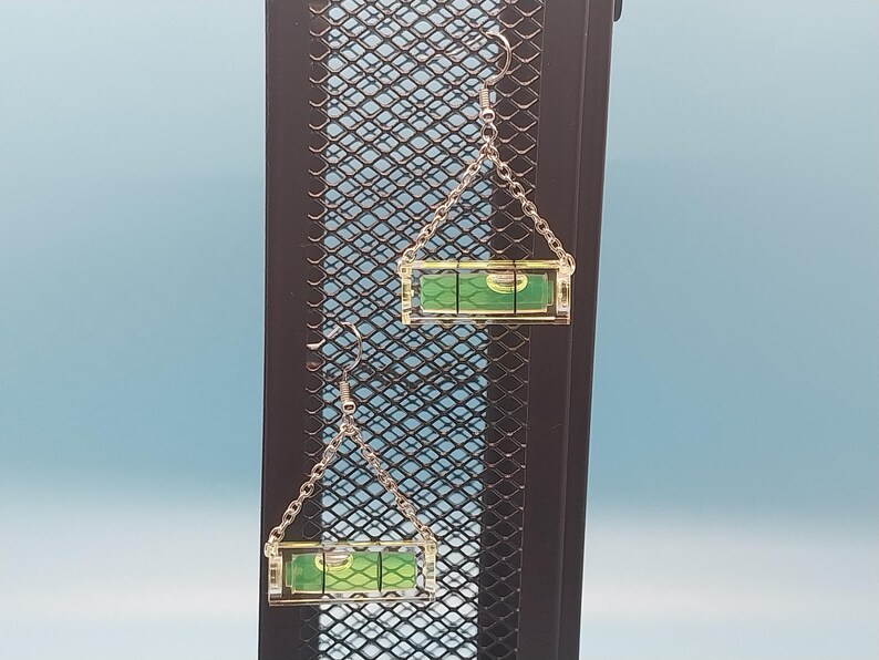 May include: A pair of novelty earrings featuring miniature levels. Each earring has a clear, rectangular level with a green liquid bubble, suspended from a silver chain and hook. The earrings are displayed against a black metal grid with a blue background.