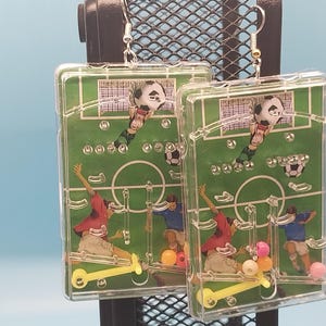May include: Clear plastic soccer-themed earrings. Each earring features a miniature soccer field design with players in red and blue uniforms, a soccer ball, and a yellow scooter. The earrings have silver-colored hooks.