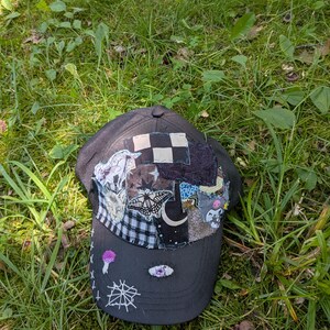 Op de afbeelding: Een zwarte baseballpet met een patchworkontwerp op het voorpaneel, bestaande uit diverse stoffen met patronen zoals ruiten en een mottenillustratie. De klep is versierd met witte geborduurde kruissteken, een spinnenweb en een klein oogmotief.