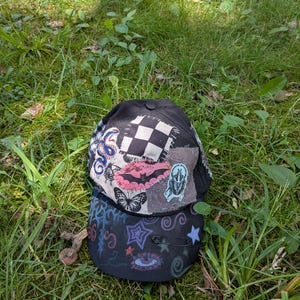 Op de afbeelding: Een zwarte baseballpet met een patchworkontwerp op de kroon, inclusief een geblokt vierkant, een roze vleermuis, een vlinder en een kever. De klep is versierd met handgetekende kleurrijke sterren, krullen en een oogmotief, met een veiligheidsspeld.