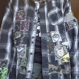 Może przedstawiać: Czarno-biała koszula w kratę z flaneli z różnymi naszywkami. Naszywki przedstawiają wzory oczu, kotów i abstrakcyjne. Koszula ma zapięcie na guziki i metalowe przelotki u dołu. Noszona na czarnym t-shircie.