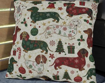 May include: A decorative square pillow with a cream background featuring a repeating pattern of dachshund dogs in festive Christmas designs. The dogs are adorned with snowflakes, ornaments, and Christmas trees. The pillow is placed on a blue cushion.